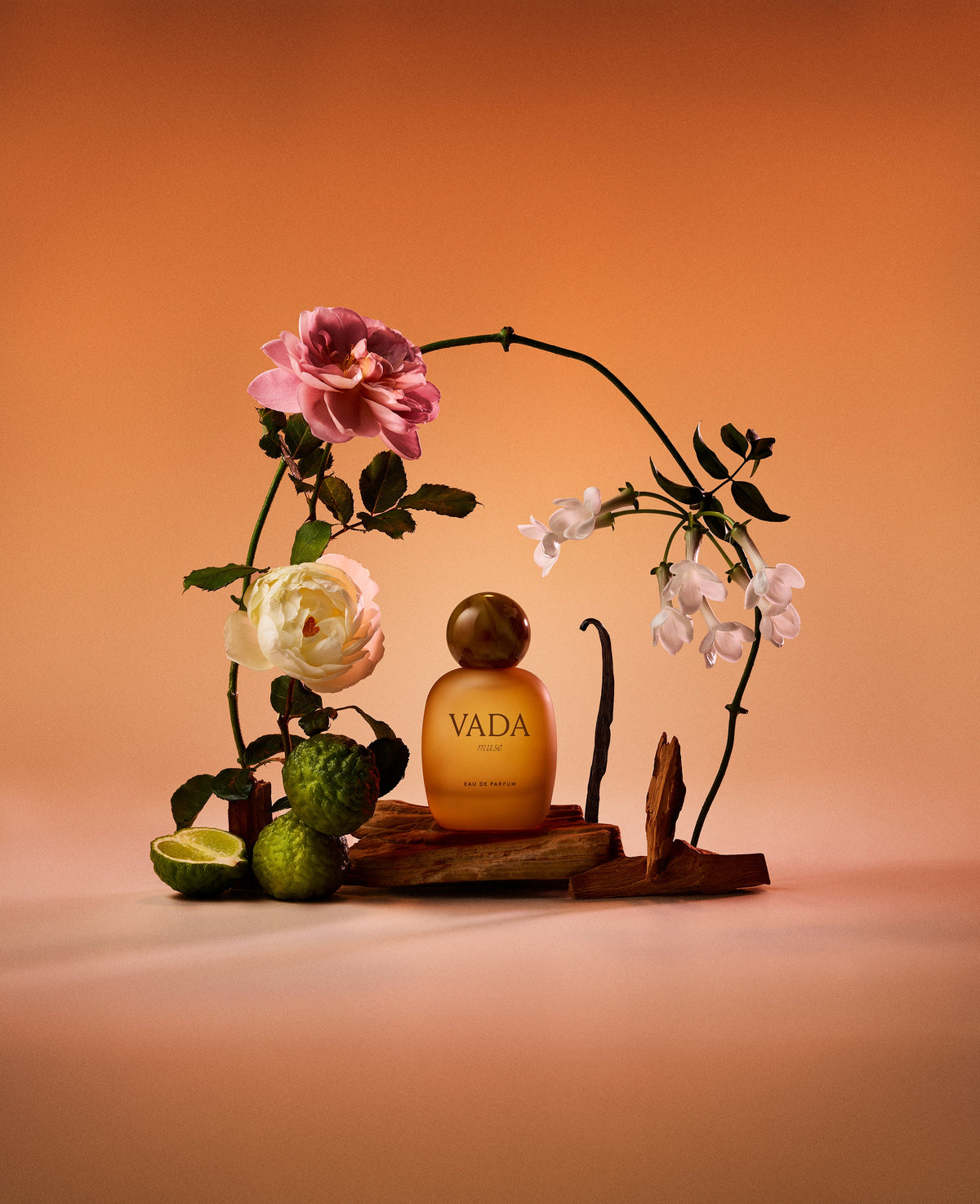 Vada perfume bottle with flowers and limes on a wooden stand against an orange background