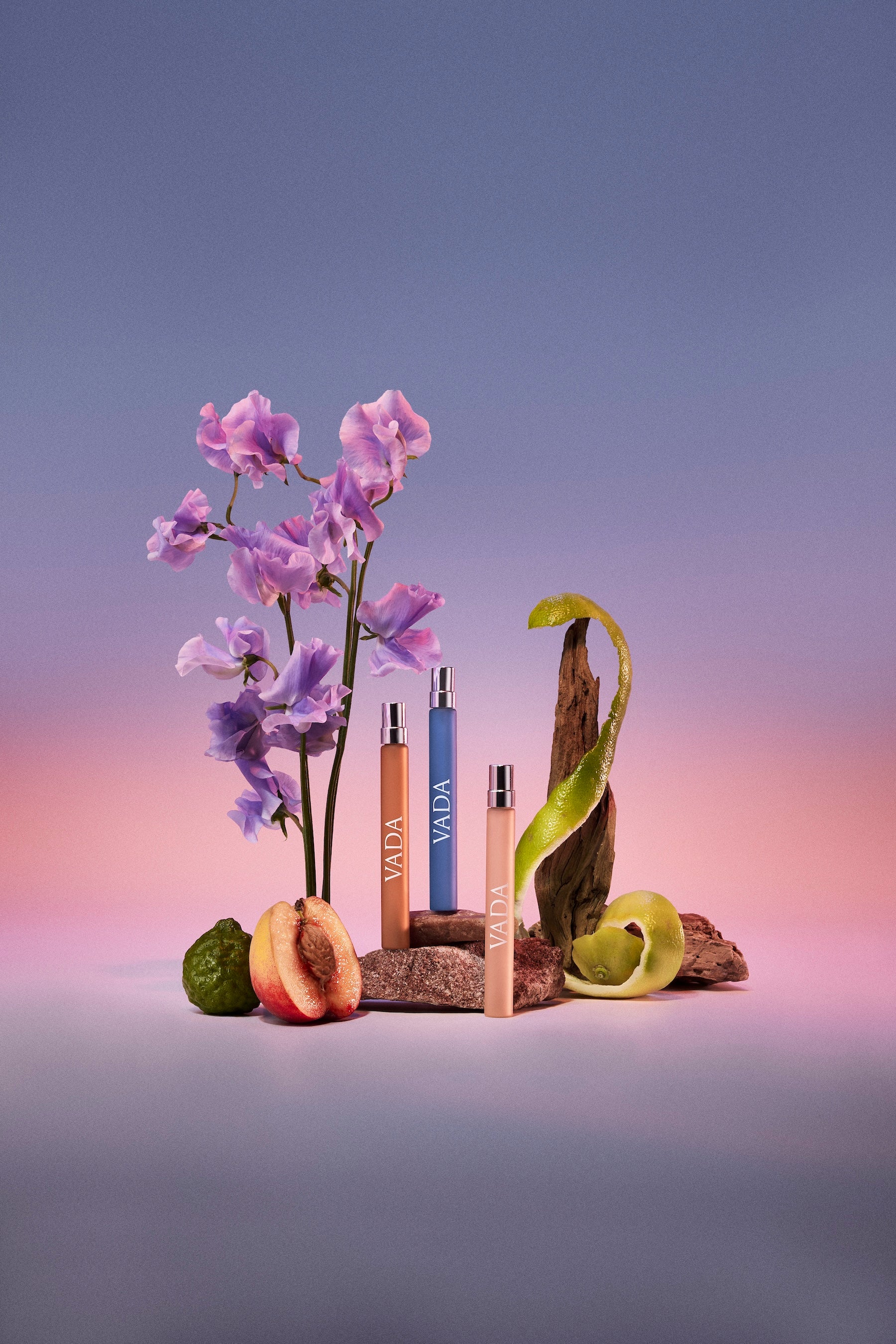 Three VADA fragrances on a wooden stand with flowers and fruits against a gradient background